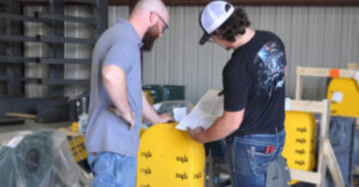 Two industrial workers reviewing technical documents next to yellow industrial equipment in a warehouse.</p>
<p>