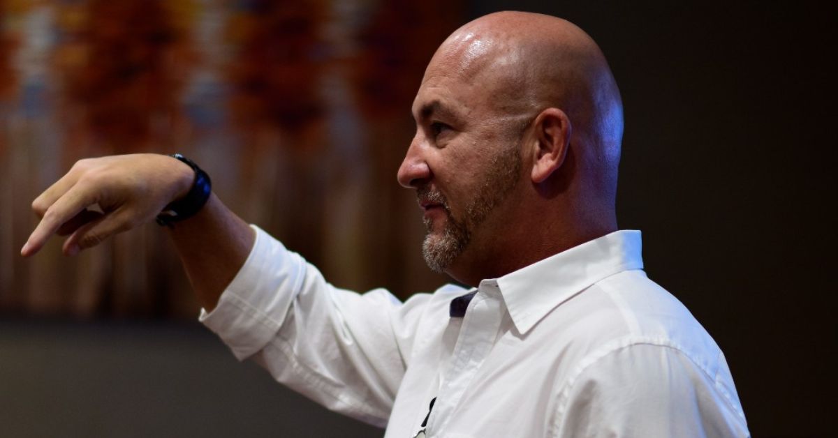 Professional speaker in a white shirt passionately delivering a presentation, gesturing towards an audience.

